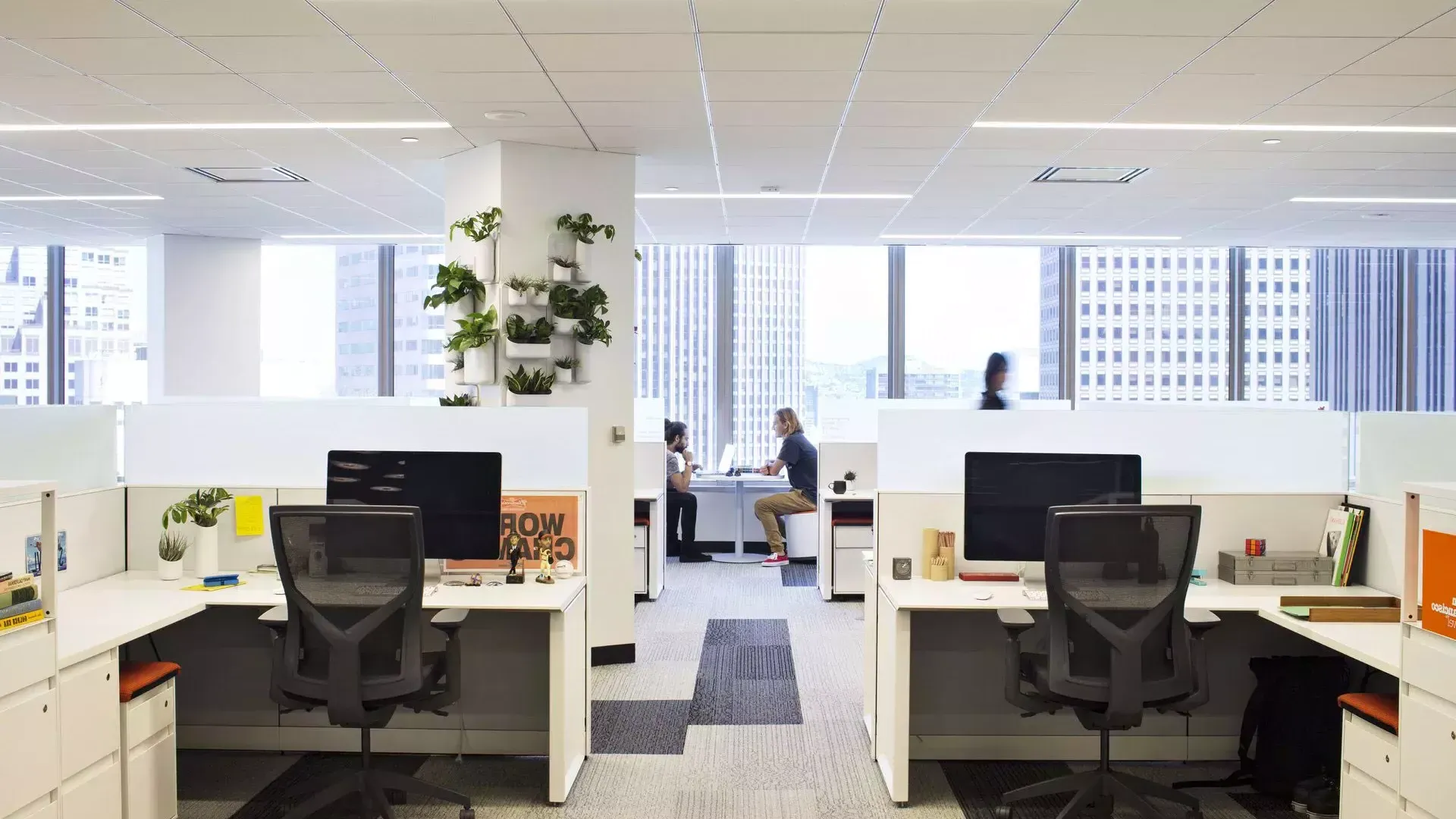 Interior scene at an office building in San Francisco, with desks in the foreground and two people working at a table in front of large windows.